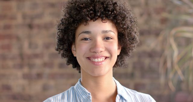 Young woman with curly hair smiling warmly. Suitable for illustrating concepts of friendliness, positivity, and confidence. Ideal for use in advertisements, online profiles, blogs, and social media content highlighting personal stories, success stories, or positive representations of everyday people.