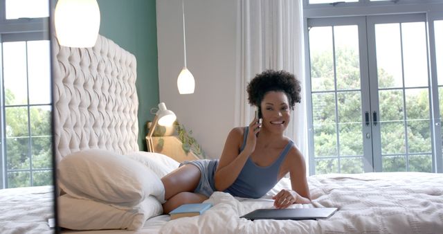 Young woman lying on bed, engaging in a casual phone conversation, with relaxed and friendly demeanor. Modern bedroom setup with natural light coming from large windows. Suitable for illustrating concepts of relaxation, casual communication, home lifestyle, comfort, and leisure.