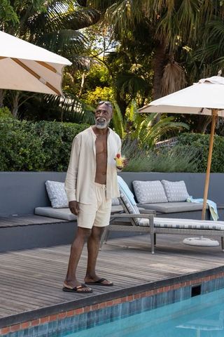 Senior african american man standing by pool holding cocktail wearing open linen shirt at tropical r