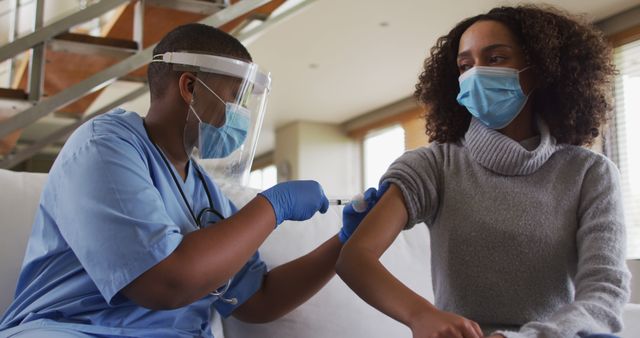 Healthcare worker in blue scrubs giving vaccine injection to masked patient on couch. Use for healthcare campaigns, vaccination promotions, medical articles, pandemic awareness.