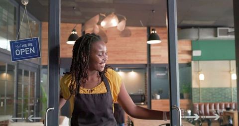Smiling cafe barista holding open glass door welcoming customers into warm coffeeshop interior