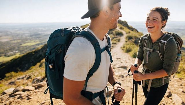 Smiling hikers trekking sunlit mountain ridge holding trekking poles and backpacks