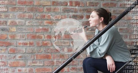 Young woman examining digital globe overlay on brick wall while using smartphone on metal stairs