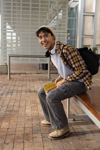 Smiling asian student sitting on campus bench holding notebook, backpack and headphones