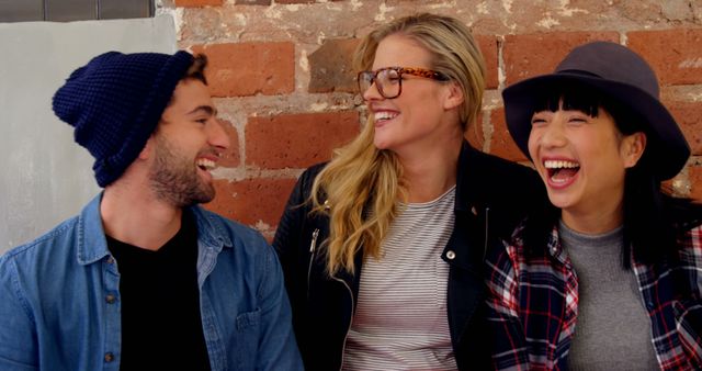 Group of young friends enjoying each other's company, laughing and smiling against a brick wall. Casual and relaxed atmosphere with diverse individuals showcasing a sense of togetherness and happiness. This image is ideal for use in promotional materials for social events, community gatherings, lifestyle blogs, fashion editorial, and positivity campaigns.