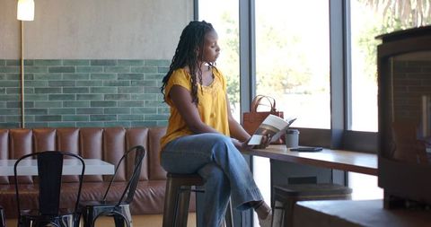 African american woman reading book at bright cafe bar wearing yellow blouse and jeans