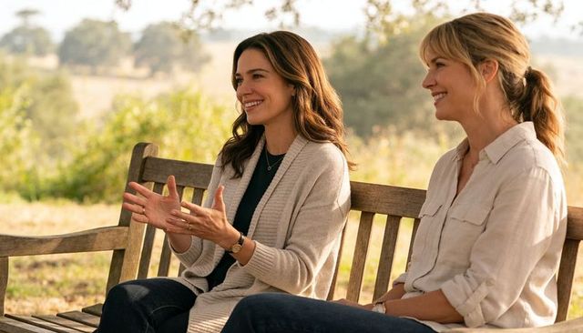 Two women smiling and conversing on wooden bench in sunlit countryside meadow