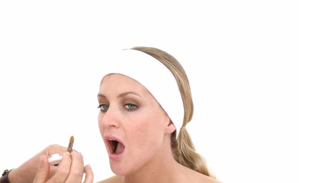 Young woman wearing white headband is applying lip gloss, focusing on personal beauty routine. Her open-mouthed expression while applying the gloss highlights attention to detail and interest in aesthetics. Useful for content related to makeup tutorials, beauty product advertisements, and skincare routines.