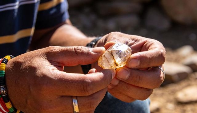 Hands holding sunlit amber crystal while examining raw gemstone outdoors