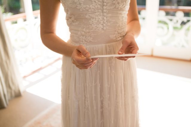 Bride standing in a sunlit room, holding a wedding card. Ideal for wedding planning websites, bridal magazines, and romantic greeting card designs.