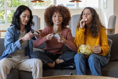 Three friends playing video games on couch while snacking and laughing together