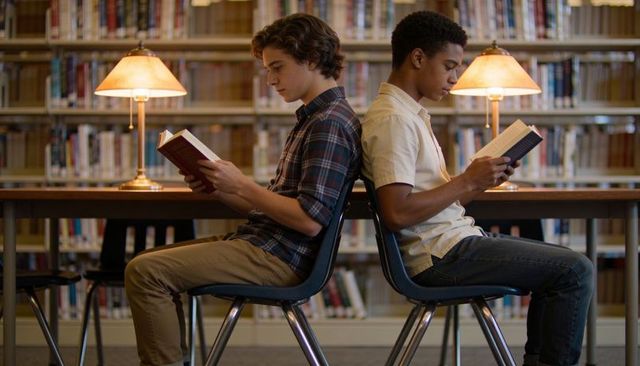 Two teen readers sitting back-to-back at library table with warm lamp glow