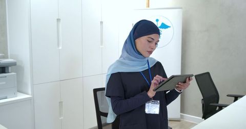 Clinic receptionist tapping tablet wearing light blue headscarf navy scrubs at front desk
