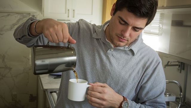 Young man pouring coffee from stainless carafe into white mug in modern marble kitchen