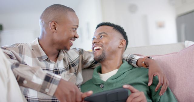 Diverse gay male couple embracing on couch and using smartphone at home. Togetherness, relationship, technology, communication and domestic life, unaltered.