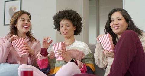 Diverse women sharing popcorn during cozy movie night on gray couch, wearing sweaters