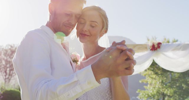 Newlywed couple sharing a romantic outdoor dance, eyes closed and smiling, under sunlight. Suitable for Wedding themes, Love and Romance advertisements, Bridal invitations, Celebrations, and Happy moments imagery.