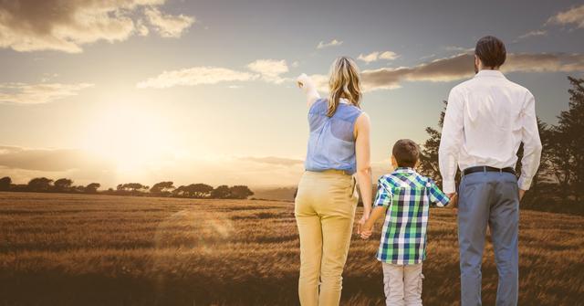 Family of three standing in open field watching sunset over countryside. Ideal for use in advertising campaigns promoting family values, outdoor activities, rural living, or travel. Perfect for websites, brochures, and posters focusing on family togetherness and nature.