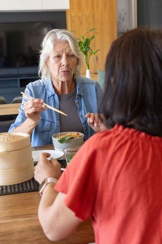 Senior women sharing noodle bowl with chopsticks at cozy dining table, smartwatch visible