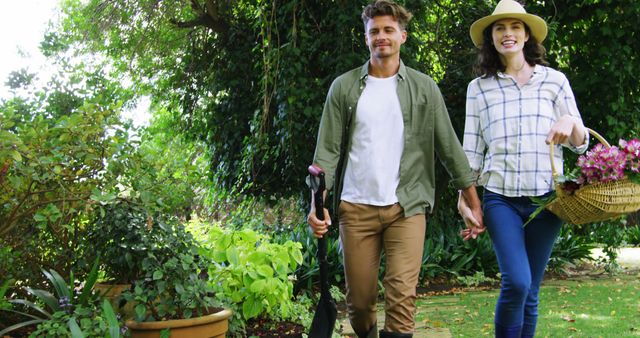 A smiling couple enjoys gardening together in a vibrant, lush garden. The woman carries a basket of flowers, while the man holds gardening tools. Ideal for concepts of teamwork, outdoor activities, gardening hobby, and spending quality time in nature.
