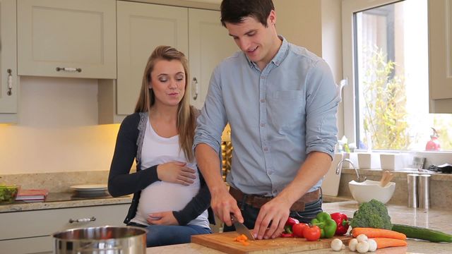 An expecting couple cooking vegetables in a bright kitchen. The man is carefully chopping vegetables while his pregnant wife gently touches her belly and watches. Ideal for themes related to family, pregnancy, healthy living, home life, and culinary activities. Can be used for websites, blogs, or campaigns focused on pregnancy, cooking, family bonding, or home lifestyle.