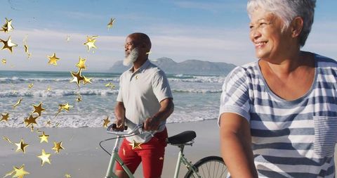 Active senior couple cycling along sunlit beach with gold star confetti celebration