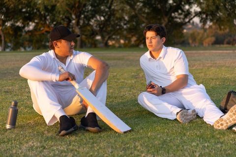 Cricket teammates resting on grass sharing equipment and conversation during golden hour