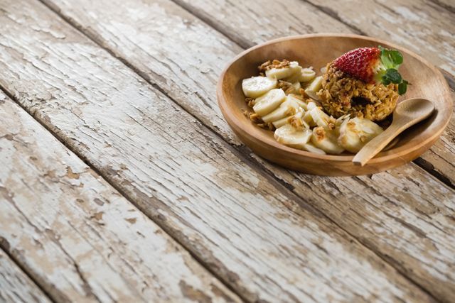 This image showcases a healthy breakfast featuring a granola bar and fresh fruits, including banana slices and a strawberry, served on a wooden plate. The rustic wooden table adds a natural and organic feel to the scene. Ideal for use in articles or advertisements related to healthy eating, breakfast recipes, nutrition, and organic food products.