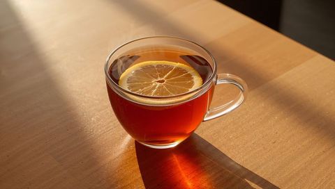 Steaming Clear Glass Cup Holding Amber Black Tea with Lemon Slice on Sunlit Wooden Table