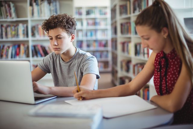 Two teenagers are studying together in a school library. One is using a laptop while the other is writing in a notebook. This image can be used for educational content, school promotions, study tips, or articles about student life and teamwork.