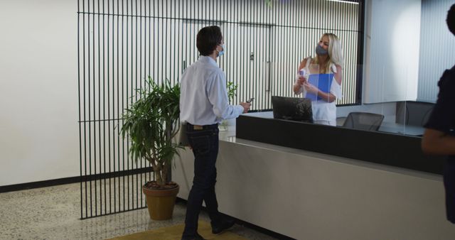 Receptionist engaging in conversation with visitor at modern office lobby front desk. Receptionist is assisting the visitor, both wearing masks, reflecting adherence to health safety protocols. The image highlights professional workspace with office furniture and decor including a planter. This can be effectively used for topics related to administrative tasks, customer service, workplace communication, health and safety adherence in office settings, and modern office environments.