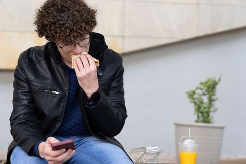 Teen male eating sandwich while checking smartphone on urban bench during lunch break