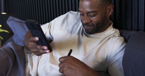 Reclining mature man holding smartphone and writing notes on bed for planning and focus