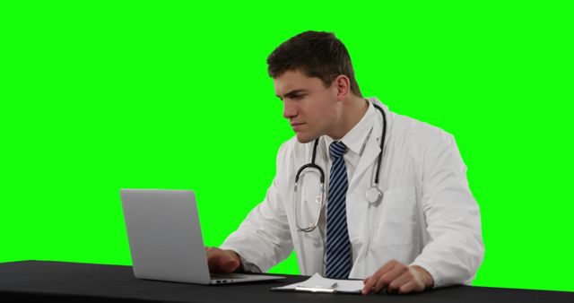 Male doctor sitting at desk, using laptop with green screen background. Suitable for healthcare, medical technology, professional presentations, or telemedicine advertisements.