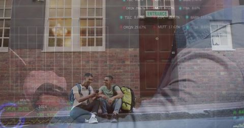 Two young men comparing smartphone on urban curb with data overlay, backpacks, headphones