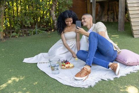 Couple enjoying backyard picnic on blanket sharing fresh fruit and taking selfie
