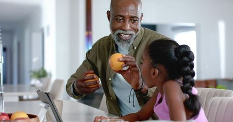 African american man offering peach to girl while using tablet in modern kitchen