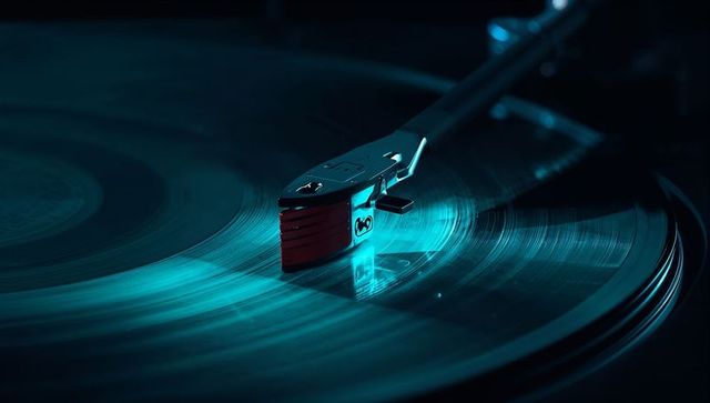 Spinning vinyl record reflecting teal light, tonearm and stylus contacting grooves