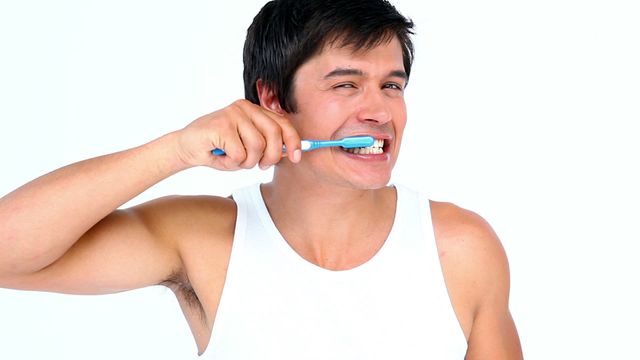 Brunette man wearing white tank top brushing teeth against a bright white setting. Ideal for use in dental hygiene promotions, health and wellness articles, personal care blog posts, and advertising oral care products.