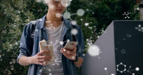 Casual young man in denim shirt checking smartphone while holding sandwich outdoors