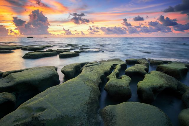 Moss-covered rock formations on a coastline during a vibrant sunset with a dramatic sky. Ideal for travel brochures, nature calendars, digital wallpapers, and social media posts. Emphasizes beauty of natural landscapes and serene evening scenes.