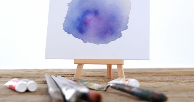 Watercolor artwork resting on small easel positioned on rustic wooden table. Surrounding it are paintbrushes, tubes of paint, and other art supplies. Ideal for illustrating creativity, artist workspaces, art studios, or DIY projects.