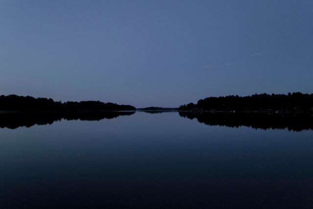 Serene twilight over a still lake with a silhouetted forested horizon. The water is calm, mirroring the sky and trees. Ideal for backgrounds, meditative content, tranquility, and nature appreciation projects.