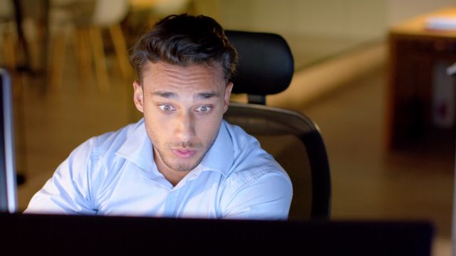 A businessman is working late at his office desk, displaying a surprised expression while using a computer. The office has a dim lighting setup, suggesting a typical corporate environment during nighttime hours. Ideal for illustrating themes related to work stress, late hours, unexpected news, or corporate lifestyle.