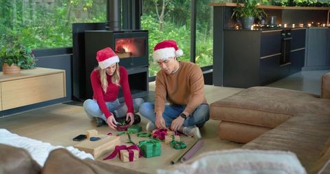 Couple wrapping christmas gifts by fireplace wearing santa hats in modern cozy living room