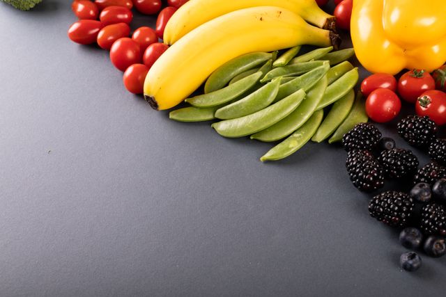 High angle view of various fresh fruits and vegetables including bananas, cherry tomatoes, snap peas, blackberries, blueberries, and a yellow bell pepper on a gray background with copy space. Ideal for use in health and wellness blogs, organic food advertisements, nutrition guides, and healthy eating campaigns.
