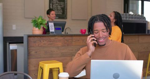 African american man working remotely in cafe using laptop and talking on phone