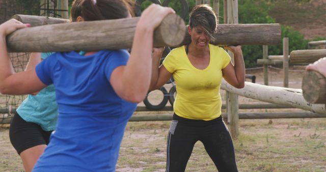 Women engaging in intense outdoor boot camp, lifting heavy wooden logs. This is a good visual for content related to fitness, group exercise, outdoor activities, strength training, and team-building exercises. Could be used in advertising for fitness programs, boot camps, athletic wear, and health and wellness articles.