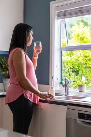 Asian woman standing at kitchen sink holding glass and pill bottle by sunny window