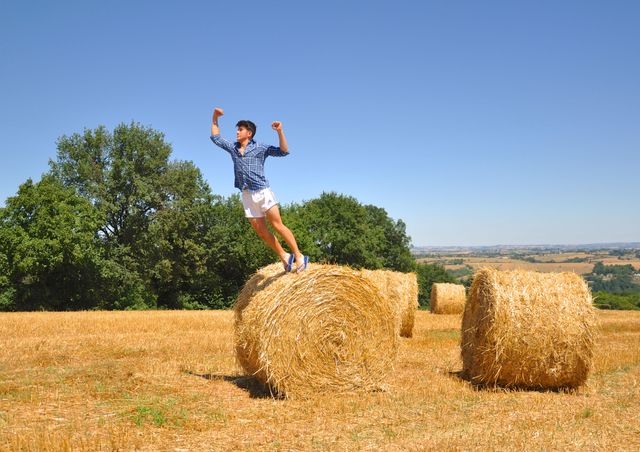 Person celebrating life and enjoying a sunny day while jumping off a hay bale. Perfect for themes related to leisure, countryside lifestyle, recreational activities, happiness in nature, and the beauty of the rural environment.